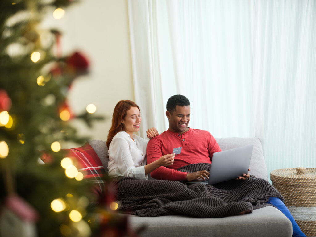 A young man using a laptop and sitting by his girlfriend that is holding a card to shop online for christmas gifts.