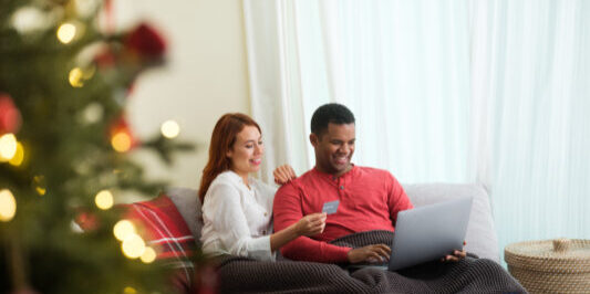 A young man using a laptop and sitting by his girlfriend that is holding a card to shop online for christmas gifts.